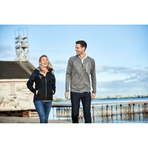 A woman wearing a black softshell jacket walks beside a man in a grey long-sleeve top by the water.