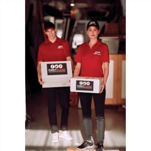 A pair of individuals wearing red polo shirts and caps, holding boxes in a warehouse setting.