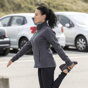 A woman stretches while wearing a grey polar fleece jacket, with parked cars in the background. The jacket features a logo.