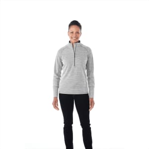 A woman wearing a grey knitted half zip jumper, smiling against a plain background. The jumper has a logo on it.