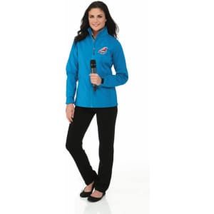 A woman in a blue softshell jacket stands with a microphone, paired with black pants and shoes. The jacket features a logo.