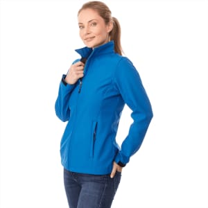 A woman wears a blue softshell jacket with a zipped collar and pockets, featuring a logo on the chest.