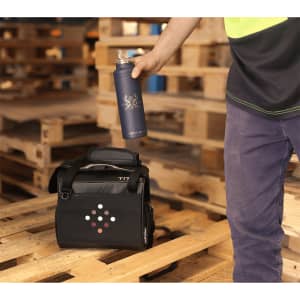 A black lunch cooler bag with a logo, beside a hand holding a blue drink bottle, set on wooden pallets.