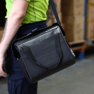 A black lunch cooler with a shoulder strap, featuring a front pocket and branding, held by a person.