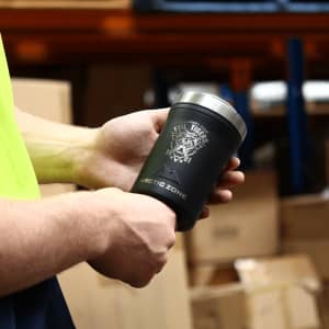 A drink bottle in black with a silver lid, featuring a logo on the side, held in hands against a warehouse backdrop.