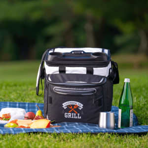 A black and white rolling cooler with a logo, placed on grass near picnic food and drinks.
