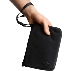 A black toiletry bag is held in a hand, featuring a zip closure and a wrist strap. It has a logo on the front.