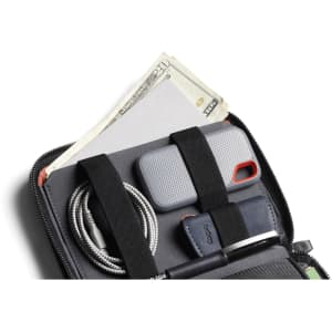 A grey toiletry bag with compartments holding cash, a phone case, a cable, and a pen against a dark background.