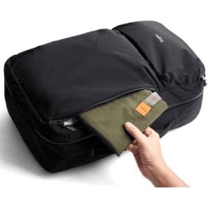 A black travel backpack with a hand reaching into a zippered pocket containing a green pouch.