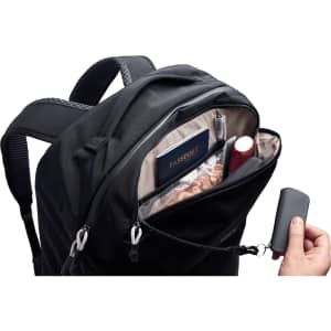 A black backpack with a zipped pocket containing a passport, pens, and a small bottle. It has a branding logo.