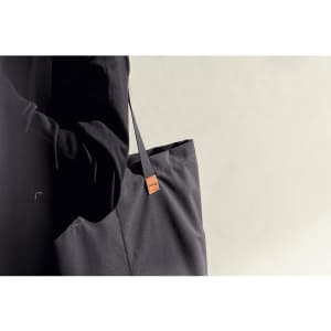 A dark grey tote bag with a branding tag, held by a person in black clothing against a neutral background.