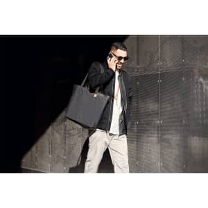 A person carrying a black tote bag, wearing sunglasses and a casual outfit, walking in a modern urban setting.