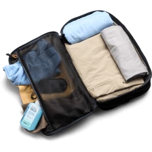 A packing cube in black, filled with rolled clothing items and a bottle of wash liquid. It has a mesh section and a logo.