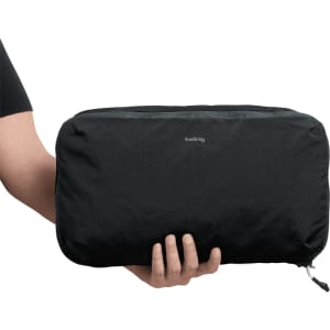 A black packing cube being held by a person, featuring a zippered closure and a small logo on the front.