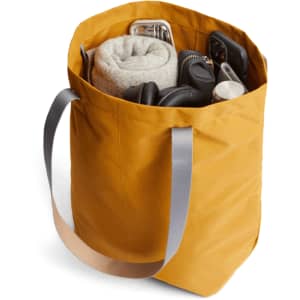 A mustard yellow tote bag filled with a blanket, headphones, and various accessories, featuring grey straps.