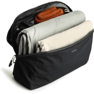 A black packing cube filled with a grey blanket, a beige item, and an orange accessory, featuring a mesh panel on top.