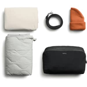 A cream blanket, grey quilted item, black packing cube, an orange beanie, and a black belt, all neatly arranged.