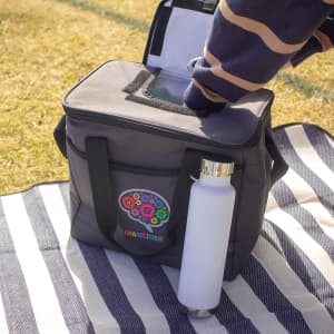 A grey cooler bag with a top opening and handles, featuring a logo, alongside a white drink bottle on a striped blanket.