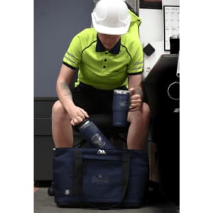 A blue drink bottle and mug sit next to a dark blue duffel bag, with a person in a high-visibility shirt and helmet.