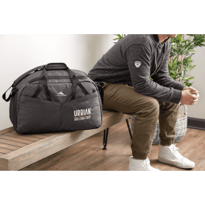 A dark grey duffel bag with a logo, placed beside a person sitting on a bench in a modern setting.