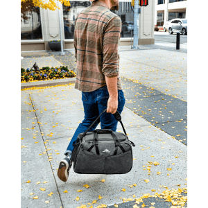 A man walks outdoors carrying a large grey duffel bag with black accents and a logo.
