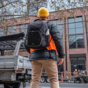 A black High Sierra Access 4.0 backpack rests on the back of a person wearing a hard hat and bright orange vest.