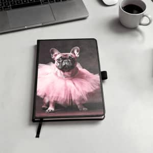 A notebook with a black cover featuring a pink tutu-wearing dog on the front. It includes a logo.