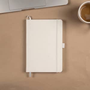 A white recycled A5 notebook with a ribbon bookmark, lying on a desk next to a laptop and a cup of coffee.