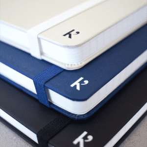 A stack of A5 hardcover notebooks in white, navy blue, and black, featuring an elastic band and a logo.