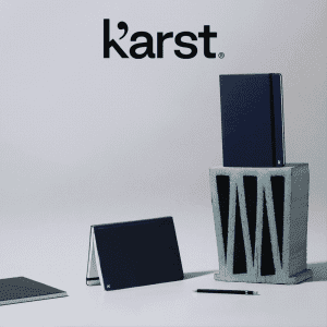 A navy hardcover notebook stands on top of a grey block, accompanied by a smaller notebook and a pen. It has a logo.