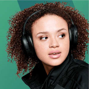 A young woman wearing black Skullcandy Hesh ANC Wireless headphones against a green background.