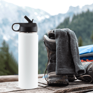 A drink bottle in white with a black lid, placed beside hiking boots and a grey scarf against a mountain backdrop.