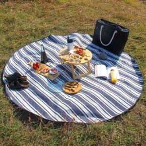A round picnic rug with navy and white stripes, featuring food, drinks, and a black tote bag on grass.