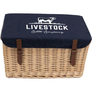 A wicker picnic basket with a navy blue canvas lid, featuring branding and leather straps for closure.