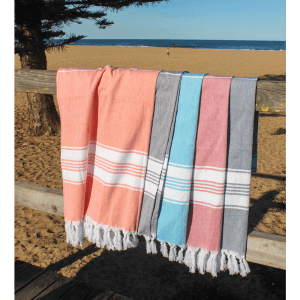 A collection of four Turkish towels in coral, blue, grey, and beige draped over a wooden railing against a beach backdrop.