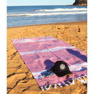 A pink and white striped Turkish towel laid on sandy beach, accompanied by a black cap and a compact camera.
