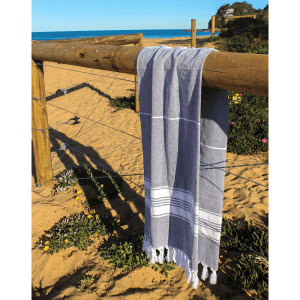 A grey and white striped Turkish towel draped over a wooden fence on a sandy beach, with ocean in the background.