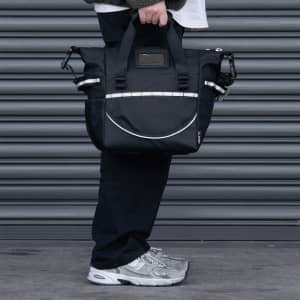 A cooler bag in black with strap and handles, featuring a logo, held by a person near a grey backdrop.