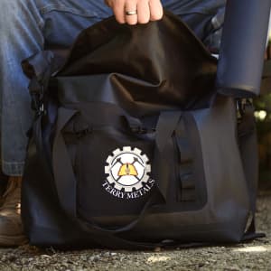 A black roll-top cooler bag with a logo, sitting beside a grey drink bottle on a textured surface.