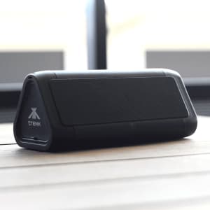 A black outdoor speaker with a triangular shape and a logo, resting on a wooden surface.