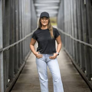 A woman wearing a black Falcon Flex Cap and a black t-shirt, standing confidently on a bridge.
