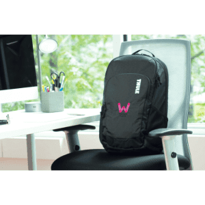 A black laptop backpack with a logo, positioned on a chair beside a desk with a computer and stationary.