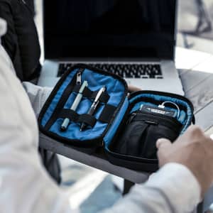 A black toiletry bag with a blue interior, containing pens and a small gadget, resting on a table beside a laptop.
