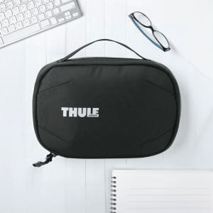 A black toiletry bag with a handle and a logo on the front, placed on a light wooden surface with office supplies.