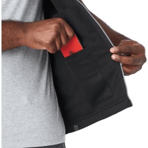 A person's hand reaching into a pocket inside a grey softshell vest, featuring a black interior. The vest has a logo.