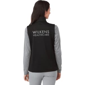 A black women's softshell vest with a high collar and grey long sleeves visible underneath, featuring a logo on the back.