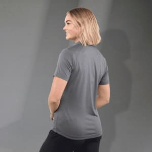 A woman wears a grey polo shirt with short sleeves, featuring a logo on the back. The background is neutral.
