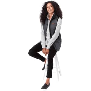 A woman sits on a stool wearing a black insulated puffer vest over a light grey top, paired with black trousers.