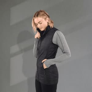 A woman wearing a black lightweight packable insulated puffer vest over a grey long-sleeve top, with hands in pockets.