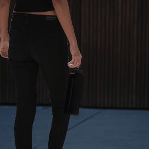 A drink bottle in matte black, held in the hand of a person wearing black jeans, standing against a textured background.